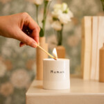 Main allumant une bougie artisanale en porcelaine blanche avec l’inscription “Maman”.
